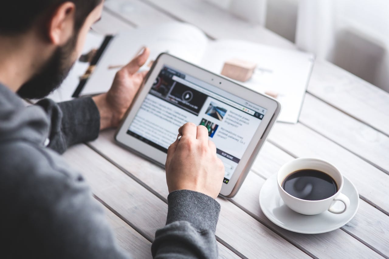 Offerings A man uses a tablet to browse news while enjoying a cup of coffee, stylus in hand.